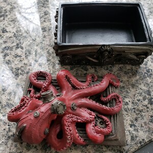 K&ouml;nnte beinhalten: Eine rote, steampunk-verzierte Oktopusfigur ist an einer Metallbox mit Zahnr&auml;dern und Zahnr&auml;dern befestigt. Die Box ist ge&ouml;ffnet und hat ein schwarzes Innenleben.