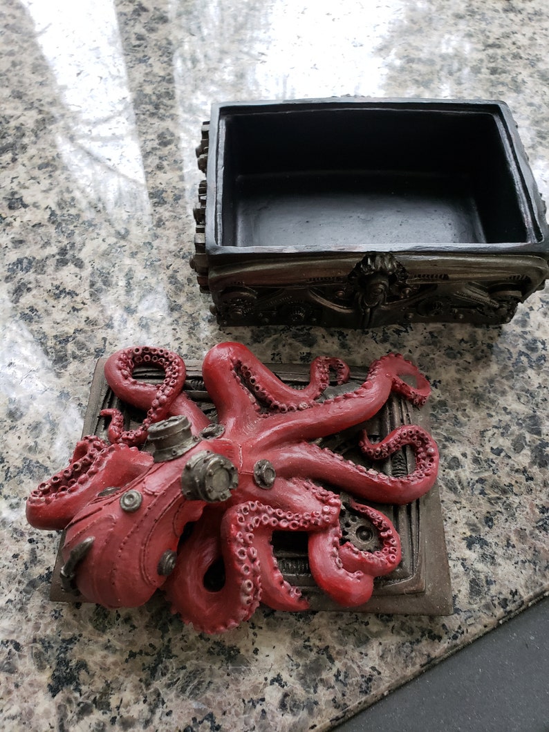 K&ouml;nnte beinhalten: Eine rote, steampunk-verzierte Oktopusfigur ist an einer Metallbox mit Zahnr&auml;dern und Zahnr&auml;dern befestigt. Die Box ist ge&ouml;ffnet und hat ein schwarzes Innenleben.