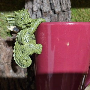Puede incluir: Una figurita de drag&oacute;n verde con un cuerpo texturizado y escamoso se aferra al lado de una taza color burdeos.