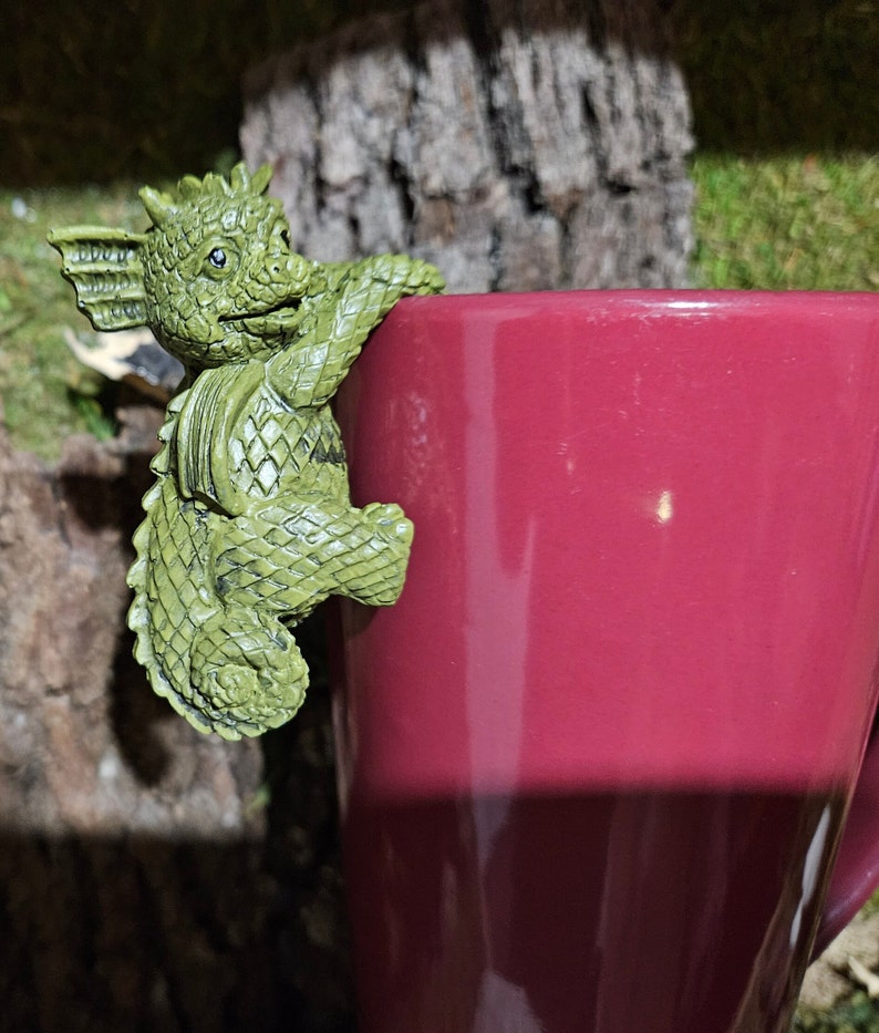 Puede incluir: Una figurita de drag&oacute;n verde con un cuerpo texturizado y escamoso se aferra al lado de una taza color burdeos.