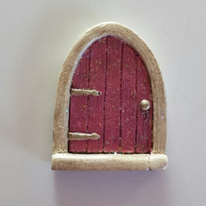 May include: A small, pink fairy door with gold accents and a white frame. The door has a rounded arch and a small knob.