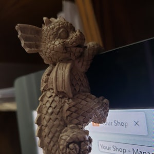 Può includere: Una piccola statuetta di drago in ceramica beige con la coda arricciata. Il drago è appollaiato sul bordo di uno schermo di computer, con la testa inclinata di lato e le ali spiegate.