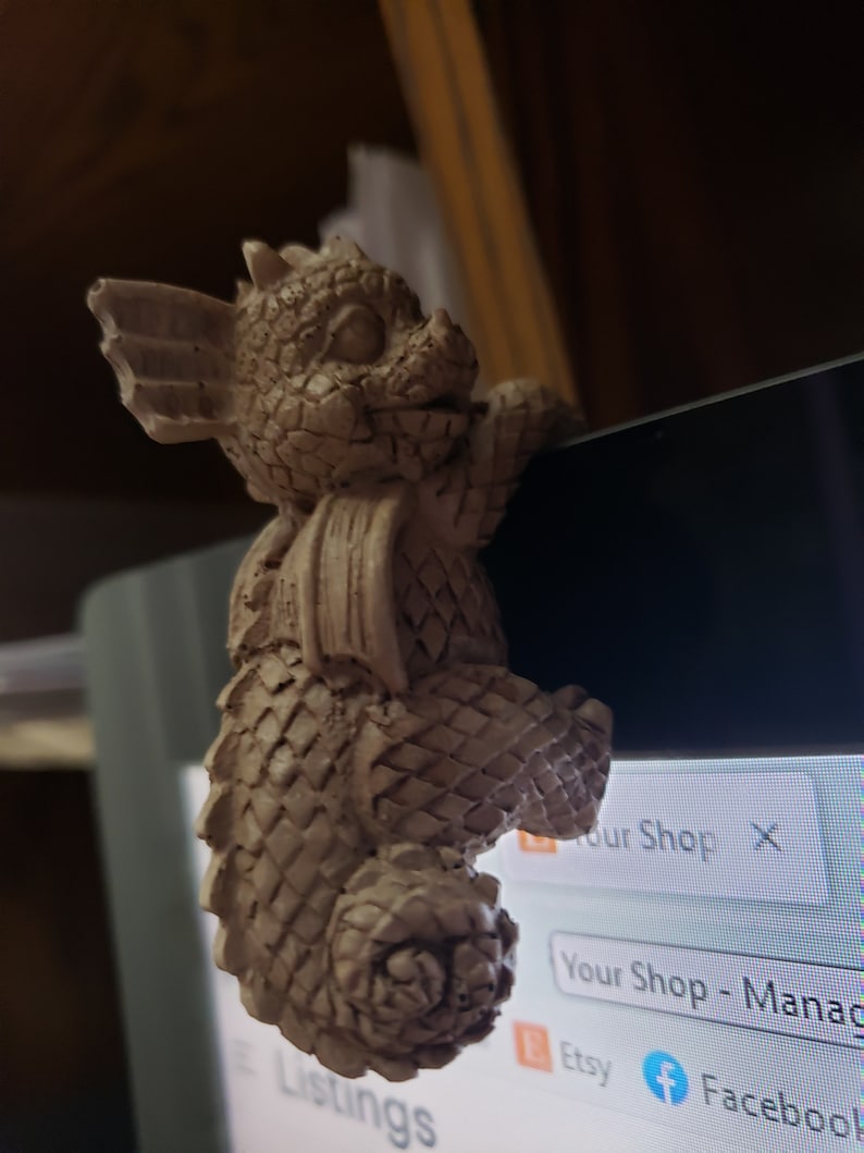 Puede incluir: Una peque&ntilde;a figura de cer&aacute;mica de drag&oacute;n beige con cola enroscada. El drag&oacute;n est&aacute; encaramado en el borde de una pantalla de ordenador, con la cabeza inclinada hacia un lado y las alas extendidas.