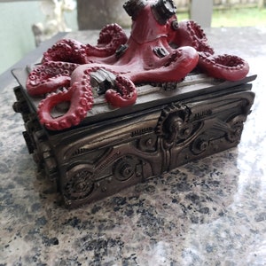 K&ouml;nnte beinhalten: Eine rote Steampunk-Oktopus-Figur mit Zahnr&auml;dern und Metallakzenten auf einer schwarzen Box mit einem Metalldesign. Der Oktopus sitzt auf der Box.