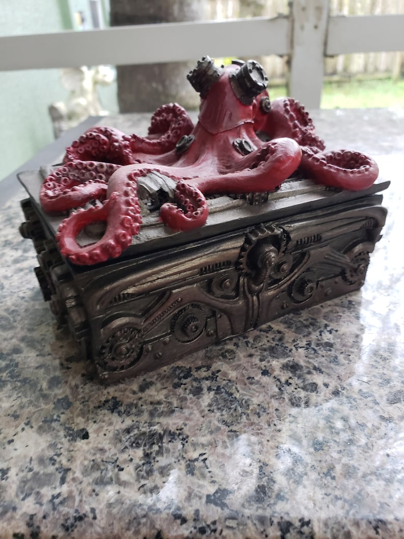 K&ouml;nnte beinhalten: Eine rote Steampunk-Oktopus-Figur mit Zahnr&auml;dern und Metallakzenten auf einer schwarzen Box mit einem Metalldesign. Der Oktopus sitzt auf der Box.