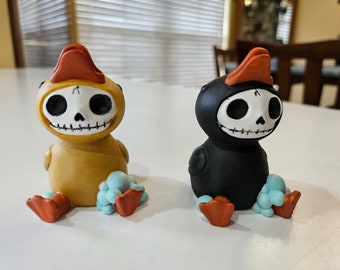 May include: Two small ceramic figurines of skeleton ducks. One is yellow and the other is black. Both have red beaks and blue feet.