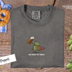 K&ouml;nnte beinhalten: Pfefferfarbenes Comfort Colors T-Shirt mit einer Cartoon-Fr&ouml;schin, die ein Bier und eine Brezel h&auml;lt, mit dem Text "WE PROST AT DAWN". Das Shirt hat einen Rundhalsausschnitt und kurze &Auml;rmel. Eine Brezel und ein Bier sind sichtbar.