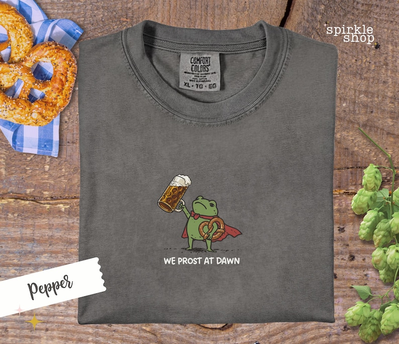 K&ouml;nnte beinhalten: Pfefferfarbenes Comfort Colors T-Shirt mit einer Cartoon-Fr&ouml;schin, die ein Bier und eine Brezel h&auml;lt, mit dem Text "WE PROST AT DAWN". Das Shirt hat einen Rundhalsausschnitt und kurze &Auml;rmel. Eine Brezel und ein Bier sind sichtbar.