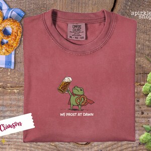 K&ouml;nnte beinhalten: Karmesinrotes T-Shirt mit einer Cartoon-Fr&ouml;schin, die einen Bierkrug und eine Brezel h&auml;lt, mit dem Text "WE PROST AT DAWN". Das Shirt hat ein Comfort Colors-Etikett. Eine Brezel und Hopfen sind im Hintergrund.