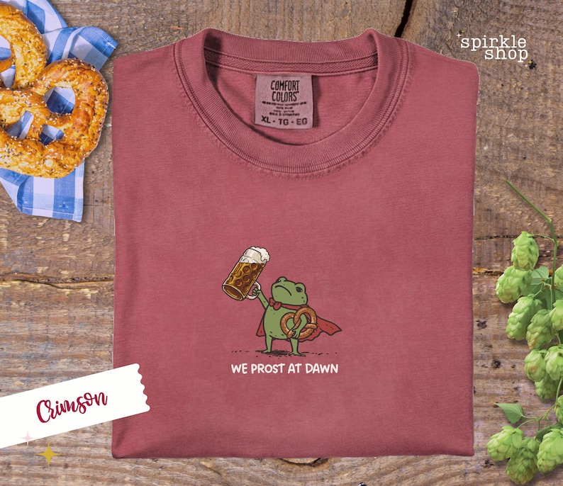 K&ouml;nnte beinhalten: Karmesinrotes T-Shirt mit einer Cartoon-Fr&ouml;schin, die einen Bierkrug und eine Brezel h&auml;lt, mit dem Text "WE PROST AT DAWN". Das Shirt hat ein Comfort Colors-Etikett. Eine Brezel und Hopfen sind im Hintergrund.