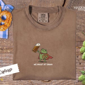 K&ouml;nnte beinhalten: Espressofarbenes T-Shirt mit einem Cartoon-Frosch, der ein Bier und eine Brezel h&auml;lt, mit dem Text "WE PROST AT DAWN". Das Shirt hat ein Comfort Colors-Etikett und liegt neben einer Brezel und Hopfen.
