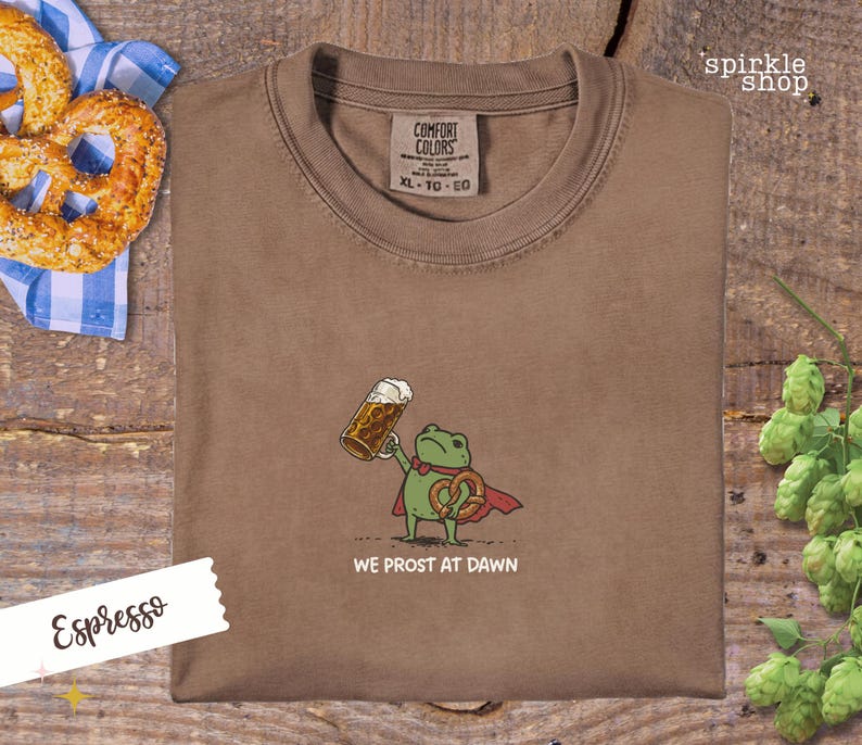 K&ouml;nnte beinhalten: Espressofarbenes T-Shirt mit einem Cartoon-Frosch, der ein Bier und eine Brezel h&auml;lt, mit dem Text "WE PROST AT DAWN". Das Shirt hat ein Comfort Colors-Etikett und liegt neben einer Brezel und Hopfen.