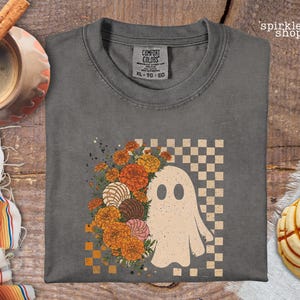 May include: A charcoal gray crewneck t-shirt featuring a graphic print of a white ghost, orange marigold flowers, and concha sweet bread pastries arranged against a beige and brown checkered background. The shirt has a Comfort Colors brand label.