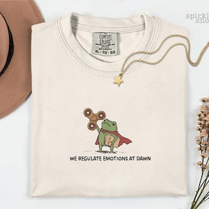 Op de afbeelding: Een cr&egrave;mekleurig t-shirt met een afbeelding van een groene kikker in een rode cape, die een bruine fidget spinner en een boek vasthoudt. De tekst onder de kikker luidt WE REGULATE EMOTIONS AT DAWN. Het shirt heeft een Comfort Colors label.