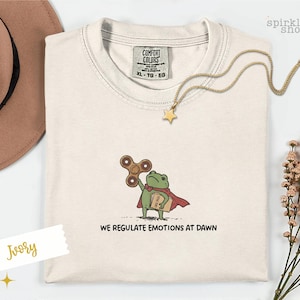 Op de afbeelding: Een ivoorkleurig t-shirt met ronde hals en een geborduurde afbeelding van een groene kikker met een rode cape, die een bruine fidget spinner en een beige boek met het label 'CASE FILES' vasthoudt. De tekst 'WE REGULATE EMOTIONS AT DAWN' staat eronder.