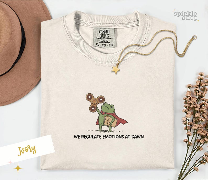 Op de afbeelding: Een ivoorkleurig t-shirt met ronde hals en een geborduurde afbeelding van een groene kikker met een rode cape, die een bruine fidget spinner en een beige boek met het label 'CASE FILES' vasthoudt. De tekst 'WE REGULATE EMOTIONS AT DAWN' staat eronder.