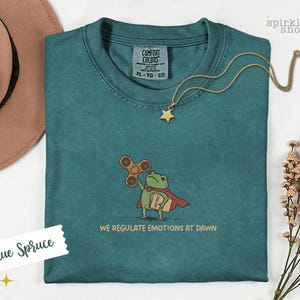 Op de afbeelding: Een blauw-sparrenkleurig t-shirt met een geborduurde afbeelding van een groene kikker in een rode cape, die een bruine fidget spinner en een beige map vasthoudt. De tekst onder de kikker luidt, WE REGULATE EMOTIONS AT DAWN. Comfort Colors label.