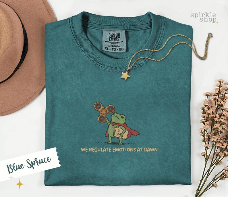 Op de afbeelding: Een blauw-sparrenkleurig t-shirt met een geborduurde afbeelding van een groene kikker in een rode cape, die een bruine fidget spinner en een beige map vasthoudt. De tekst onder de kikker luidt, WE REGULATE EMOTIONS AT DAWN. Comfort Colors label.
