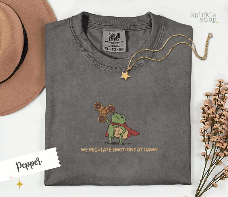 Op de afbeelding: Een antracietgrijs t-shirt met een borduursel van een groene kikker in een rode cape, die een bruine fidget spinner en een beige map met het opschrift CASE FILES vasthoudt. De tekst eronder luidt WE REGULATE EMOTIONS AT DAWN.