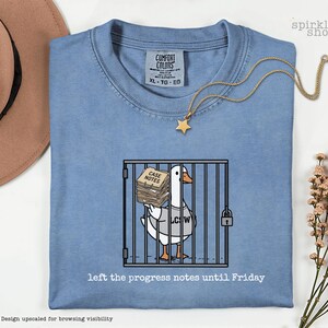 May include: A blue t-shirt featuring a graphic of a white goose inside a cage, holding a stack of papers labeled "CASE NOTES" and wearing a shirt that says "LCSW". Below the graphic, the text reads "left the progress notes until Friday".