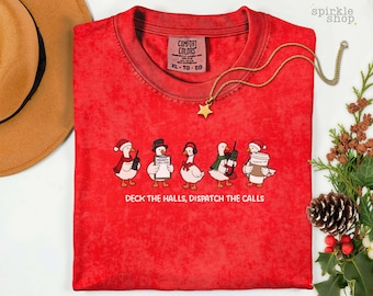 Op de afbeelding: Een rood t-shirt met een afbeelding van vijf ganzen in feestelijke kleding, waaronder kerstmutsen en sjaals, die portofoons, een klembord en een koffiebeker vasthouden. De tekst luidt: "DECK THE HALLS, DISPATCH THE CALLS."