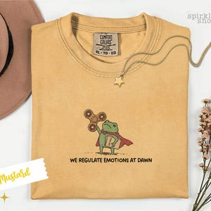 Op de afbeelding: Een mosterdgeel t-shirt met een geborduurde afbeelding van een groene kikker in een rode cape, die een bruine fidget spinner en een boek vasthoudt. De tekst onder de kikker luidt "WE REGULATE EMOTIONS AT DAWN". Het shirt heeft een Comfort Colors label.