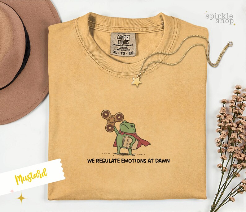 Op de afbeelding: Een mosterdgeel t-shirt met een geborduurde afbeelding van een groene kikker in een rode cape, die een bruine fidget spinner en een boek vasthoudt. De tekst onder de kikker luidt "WE REGULATE EMOTIONS AT DAWN". Het shirt heeft een Comfort Colors label.