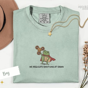 Op de afbeelding: Een lichtgroen t-shirt met een geborduurde afbeelding van een groene kikker in een rode cape, die een bruine fidget spinner en een beige map met het opschrift "CASE FILES" vasthoudt. De tekst "WE REGULATE EMOTIONS AT DAWN" staat in het zwart eronder.