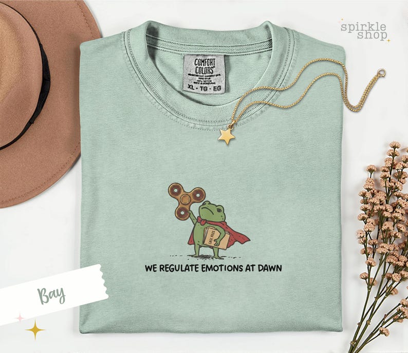 Op de afbeelding: Een lichtgroen t-shirt met een geborduurde afbeelding van een groene kikker in een rode cape, die een bruine fidget spinner en een beige map met het opschrift "CASE FILES" vasthoudt. De tekst "WE REGULATE EMOTIONS AT DAWN" staat in het zwart eronder.