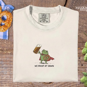 K&ouml;nnte beinhalten: Cremefarbenes T-Shirt mit einer Cartoon-Fr&ouml;schin, die einen Bierkrug und eine Brezel h&auml;lt. Der Frosch tr&auml;gt einen roten Umhang und der Text "WE PROST AT DAWN" steht darunter. Eine Brezel und eine blau-wei&szlig; karierte Serviette sind im Hintergrund.