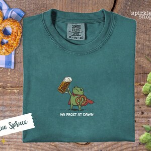 K&ouml;nnte beinhalten: Blaugr&uuml;nes T-Shirt mit einer Cartoon-Fr&ouml;schin, die ein Bier und eine Brezel h&auml;lt und einen roten Umhang tr&auml;gt. Der Text "WE PROST AT DAWN" steht darunter. Das Shirt hat ein Comfort Colors-Etikett.