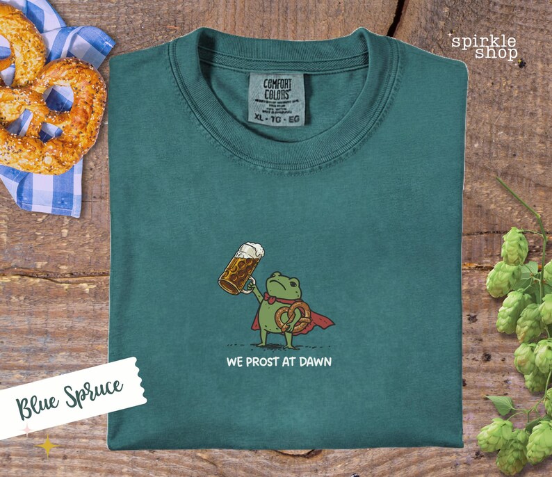 K&ouml;nnte beinhalten: Blaugr&uuml;nes T-Shirt mit einer Cartoon-Fr&ouml;schin, die ein Bier und eine Brezel h&auml;lt und einen roten Umhang tr&auml;gt. Der Text "WE PROST AT DAWN" steht darunter. Das Shirt hat ein Comfort Colors-Etikett.