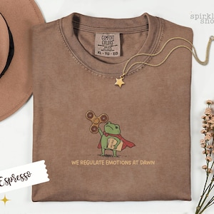 Op de afbeelding: Een espresso-kleurig t-shirt met een geborduurde afbeelding van een groene kikker in een rode cape, die een bruine fidget spinner en een beige map met het opschrift "CASE FILES" vasthoudt. De tekst "WE REGULATE EMOTIONS AT DAWN" staat eronder.