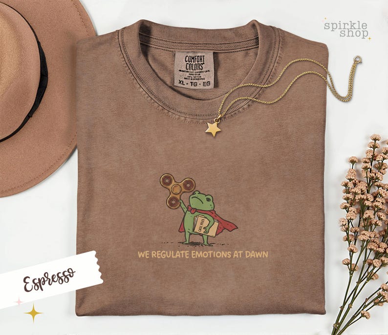 Op de afbeelding: Een espresso-kleurig t-shirt met een geborduurde afbeelding van een groene kikker in een rode cape, die een bruine fidget spinner en een beige map met het opschrift "CASE FILES" vasthoudt. De tekst "WE REGULATE EMOTIONS AT DAWN" staat eronder.