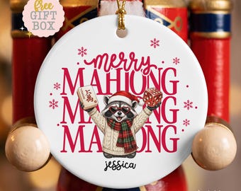 Op de afbeelding: Een rond wit keramieken ornament met een wasbeer die een kerstmuts, hartvormige zonnebril en een geruite sjaal draagt. De wasbeer houdt een mahjongtegel en een rode dobbelsteen vast. Tekst merry MAHJONG MAHJONG en jessica, met sneeuwvlokken.