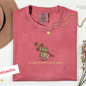 Op de afbeelding: Een watermeloenkleurig t-shirt met een geborduurde groene kikker in een rode cape, die een bruine fidget spinner en een boek vasthoudt. Onder de kikker staat de tekst WE REGULATE EMOTIONS AT DAWN. Het shirt heeft een Comfort Colors label in maat XL.