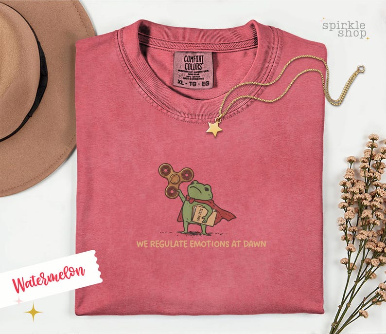 Op de afbeelding: Een watermeloenkleurig t-shirt met een geborduurde groene kikker in een rode cape, die een bruine fidget spinner en een boek vasthoudt. Onder de kikker staat de tekst WE REGULATE EMOTIONS AT DAWN. Het shirt heeft een Comfort Colors label in maat XL.