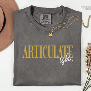 May include: A charcoal gray crew neck t-shirt with the text ARTICULATE in gold and ish. in white printed on the front. The shirt features a Comfort Colors label in size XL and is displayed with a tan hat, a gold star necklace, and dried flowers.