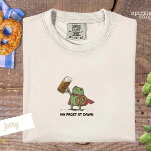 K&ouml;nnte beinhalten: Elfenbeinfarbenes T-Shirt mit einem Cartoon-Frosch, der ein Bier und eine Brezel h&auml;lt und einen Umhang tr&auml;gt. Der Text "WE PROST AT DAWN" steht darunter. Eine Brezel und eine blau-wei&szlig; karierte Serviette sind im Hintergrund.