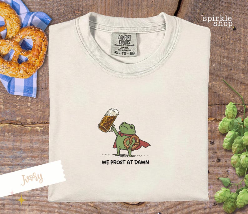 K&ouml;nnte beinhalten: Elfenbeinfarbenes T-Shirt mit einem Cartoon-Frosch, der ein Bier und eine Brezel h&auml;lt und einen Umhang tr&auml;gt. Der Text "WE PROST AT DAWN" steht darunter. Eine Brezel und eine blau-wei&szlig; karierte Serviette sind im Hintergrund.