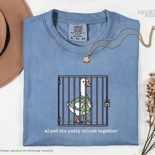 May include: A blue t-shirt featuring a graphic of a white goose inside a metal cage, holding a glob of multicolored putty. The text below the cage reads, "mixed the putty colors together." The shirt has a Comfort Colors label in size XL.