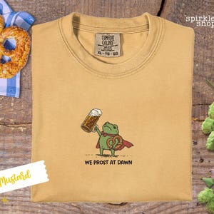 K&ouml;nnte beinhalten: Senffarbenes T-Shirt mit einem Cartoon-Frosch, der einen Bierkrug und eine Brezel h&auml;lt. Der Frosch tr&auml;gt einen roten Umhang und der Text "WE PROST AT DAWN" steht darunter. Das Shirt hat ein Comfort Colors-Etikett.