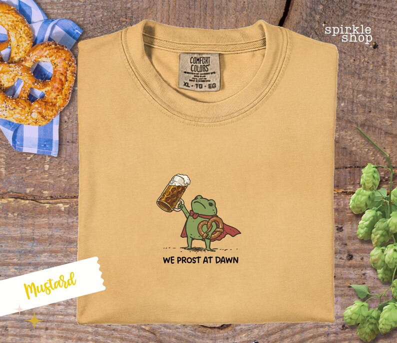 K&ouml;nnte beinhalten: Senffarbenes T-Shirt mit einem Cartoon-Frosch, der einen Bierkrug und eine Brezel h&auml;lt. Der Frosch tr&auml;gt einen roten Umhang und der Text "WE PROST AT DAWN" steht darunter. Das Shirt hat ein Comfort Colors-Etikett.