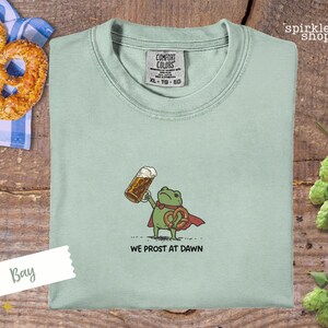 K&ouml;nnte beinhalten: Hellgr&uuml;nes T-Shirt mit einem Cartoon-Frosch, der ein Bier und eine Brezel h&auml;lt. Der Frosch tr&auml;gt einen roten Umhang und der Text "WE PROST AT DAWN" steht darunter. Das Shirt hat ein Comfort Colors-Etikett.