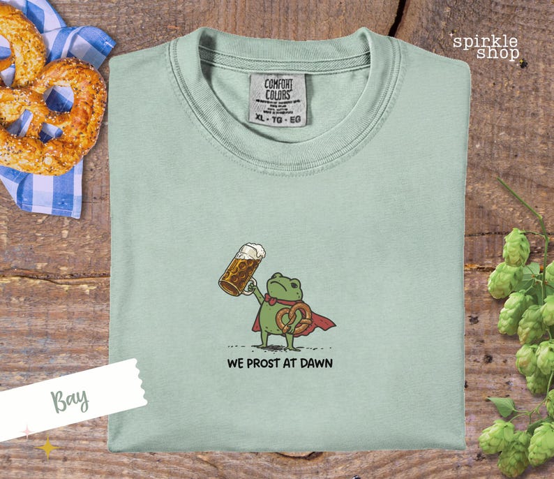 K&ouml;nnte beinhalten: Hellgr&uuml;nes T-Shirt mit einem Cartoon-Frosch, der ein Bier und eine Brezel h&auml;lt. Der Frosch tr&auml;gt einen roten Umhang und der Text "WE PROST AT DAWN" steht darunter. Das Shirt hat ein Comfort Colors-Etikett.