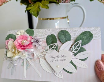 May include: A rectangular wedding invitation card featuring a white heart-shaped cutout with the text Doug & Pam 12-13-2026. The card is decorated with layered paper flowers in pink and cream, green paper leaves, white die-cut foliage, and a white ribbon bow.