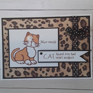 May include: A handmade greeting card with a cartoon cat illustration. The cat is orange and white, with the text "Meo-owch!" and "CAT heard you had -aract surgery". The background features a leopard print design and a black bow.