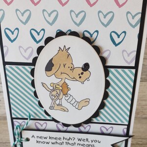 May include: Greeting card with a cartoon dog illustration. The dog has a bandage on its leg. The card has a white background with pink, teal, and purple hearts. The text reads: "A new knee huh? Well, you know what that means..."
