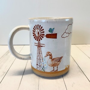 May include: White ceramic mug featuring a brown windmill, a duck, a butterfly, and clouds. The mug has a brown rim and a brown base.