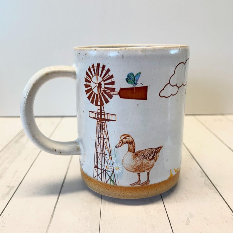 May include: White ceramic mug featuring a brown windmill, a duck, a butterfly, and clouds. The mug has a brown rim and a brown base.